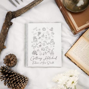 May include: A white spiral-bound notebook with the text "Getting Hitched Future Mrs. Smith" and wedding-themed doodles. The image includes a pine cone, flowers, and a vintage book.