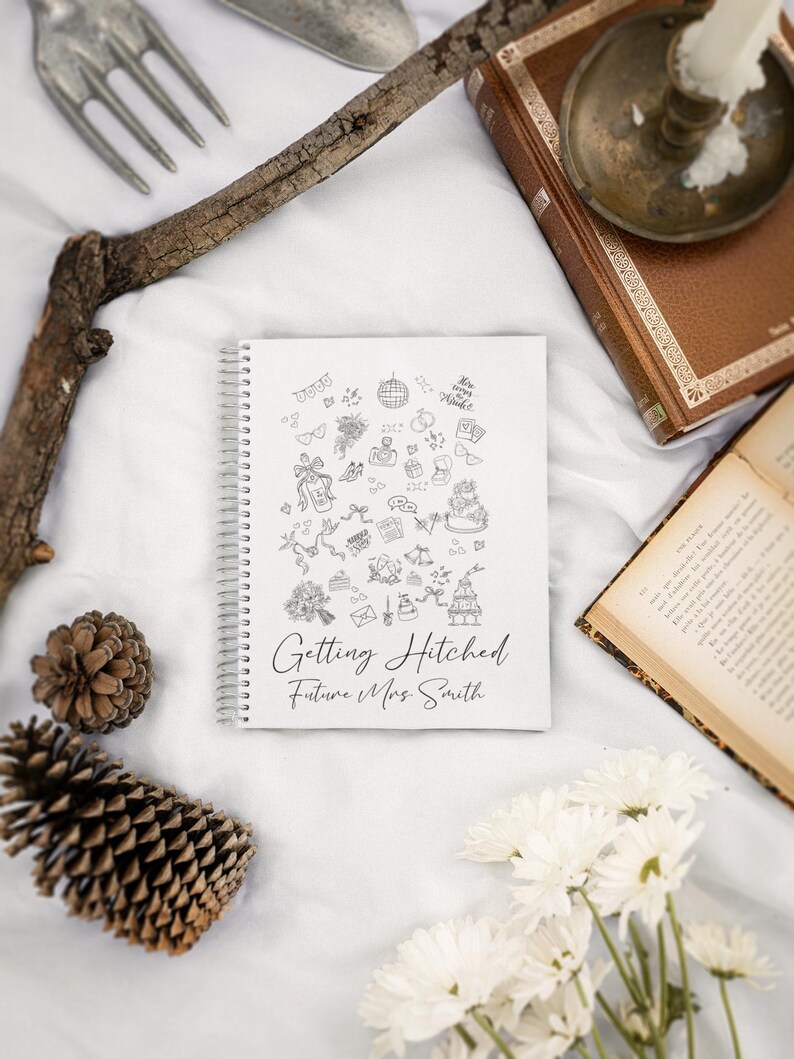 May include: A white spiral-bound notebook with the text "Getting Hitched Future Mrs. Smith" and wedding-themed doodles. The image includes a pine cone, flowers, and a vintage book.