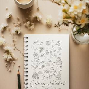 May include: A spiral-bound notebook with the words "Getting Hitched Future Mrs. Smith" and wedding-themed doodles. A cup of tea, flowers, and a pencil are arranged around the notebook on a beige surface.