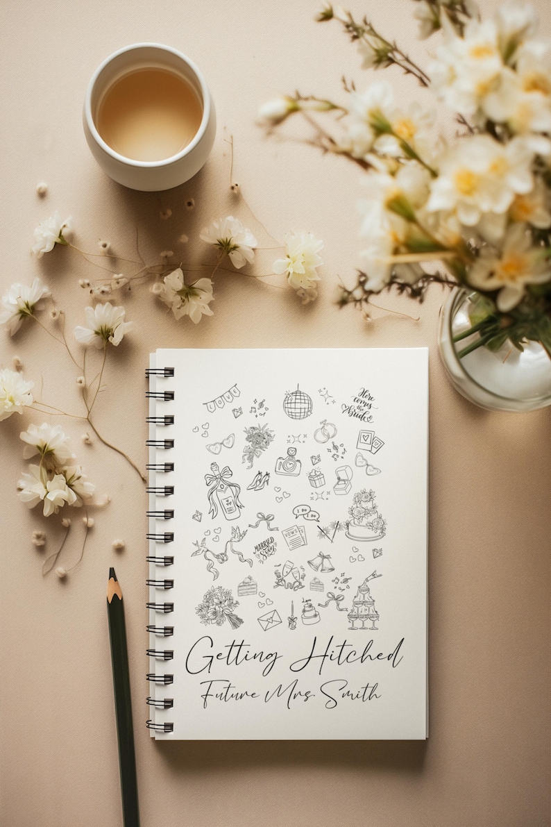 May include: A spiral-bound notebook with the words "Getting Hitched Future Mrs. Smith" and wedding-themed doodles. A cup of tea, flowers, and a pencil are arranged around the notebook on a beige surface.