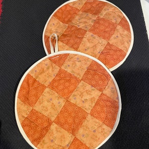 May include: Two round quilted pot holders with a white border. The pot holders have a checkered pattern in shades of orange and brown with a floral print.