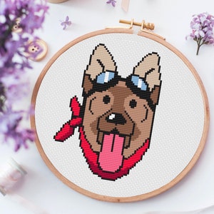 May include: Cross-stitch embroidery hoop with a pixelated image of a brown dog wearing blue goggles and a red bandana. The dog is sticking its tongue out.