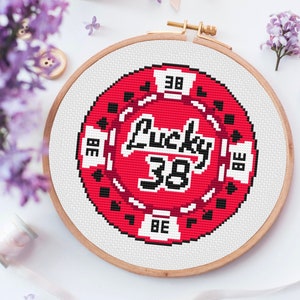 May include: A red and black cross-stitch embroidery hoop with the text "Lucky 38" in white. The hoop is on a white surface.