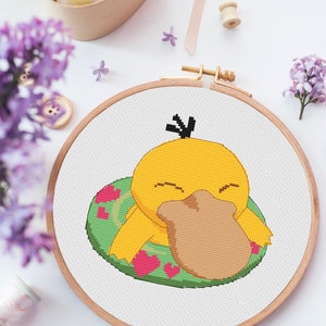May include: A completed cross-stitch embroidery in a wooden hoop. The design features a yellow cartoon duck with a brown bill, resting on a green float with pink hearts. The background is white, with lilac flowers and sewing supplies.