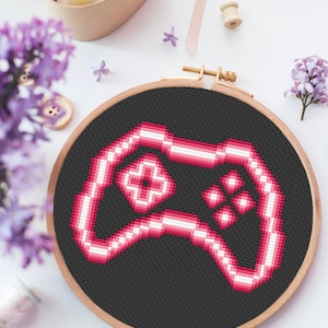 Puede incluir: Un diseño de mando de videojuego pixelado rosa y blanco sobre tela negra en un aro de bordado de madera.