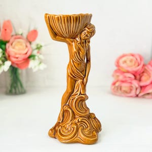 Peut inclure: Une figurine en céramique marron brillant représentant une personne debout sur une base tourbillonnante, tenant un grand bol cannelé en forme de coquillage au-dessus de sa tête. La silhouette est drapée et la base présente des détails floraux.