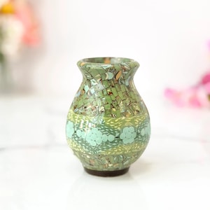 May include: A small, decorative vase with a mosaic design. The vase features a green and tan color scheme with a band of light blue floral accents. The vase has a rounded body and a slightly flared opening. The base is a dark brown color.