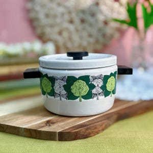 May include: A white ceramic pot with a lid and black handles. The pot is decorated with a repeating pattern of green and white stylized trees. The pot is sitting on a wooden cutting board.