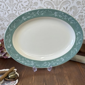 Könnte beinhalten: Eine ovale Servierplatte mit weißer Mitte und türkisfarbenem Rand. Der Rand ist mit einem zarten weißen Blumen- und Blättermuster verziert. Die Platte wird auf einem transparenten Ständer präsentiert.