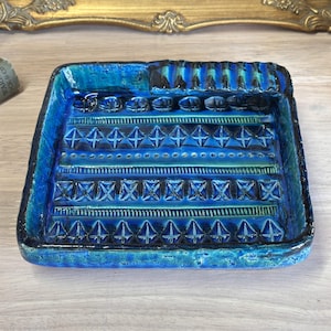May include: A rectangular ceramic ashtray with a striking blue glaze. The ashtray has a textured surface with geometric patterns and a raised edge. The design incorporates shades of turquoise, teal, and black, creating a visual contrast. The ashtray measures approximately 15 cm by 15 cm.