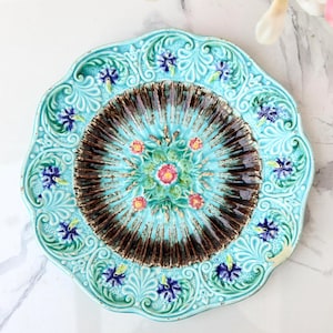 Puede incluir: Un plato de cerámica redondo con borde festoneado y esmalte turquesa. El centro presenta un patrón estriado radiante de color marrón oscuro alrededor de un motivo floral central con acentos rosas y verdes, rodeado de pequeñas flores azules.