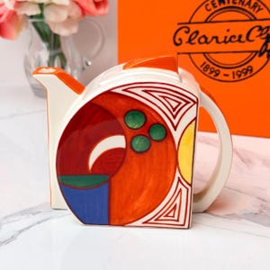 Può includere: Una teiera in ceramica con un design geometrico dai colori vivaci come arancione, rosso, blu, giallo e verde. La teiera presenta un manico e un beccuccio bianchi, una parte superiore piatta e un pannello laterale curvo decorato con forme astratte e tre cerchi verdi.