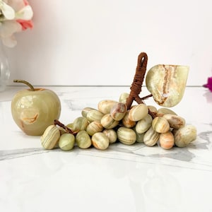 May include: A polished light green stone apple figurine next to a cluster of individual light green stone grapes. The grapes are connected by a brown wrapped stem and feature a single carved stone leaf attached to the top of the cluster.