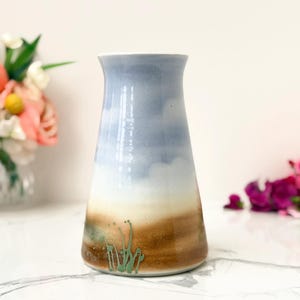 May include: A ceramic vase with a tapered cylindrical shape featuring a gradient color scheme. The top section is a soft sky blue, transitioning into a creamy white middle, and ending in a warm brown base with green painted grass details.