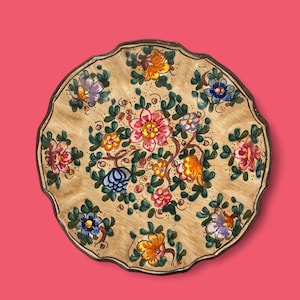 Puede incluir: Un plato decorativo de cerámica con borde festoneado, con fondo beige y diseño floral. Las flores son en tonos rosa, naranja, azul y morado, con hojas verdes y un borde marrón oscuro. El plato tiene un diámetro de aproximadamente 25 cm.