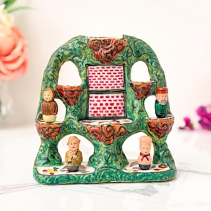 May include: A green ceramic display stand with a textured, vine-like surface and two central rectangular panels featuring a red and white brick pattern. Four small, hand-painted figurines are positioned on the integrated shelves of the structure.