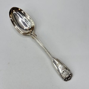 Puede incluir: Una cuchara plateada con un cuenco ovalado y un mango largo y acanalado. El mango tiene un diseño decorativo de concha. La cuchara está hecha de un metal brillante y reflectante.