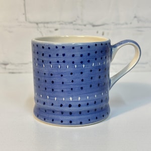 May include: A blue ceramic mug with a white interior and handle. The mug features a pattern of dark blue and white dots. The cylindrical mug has a slightly flared base.