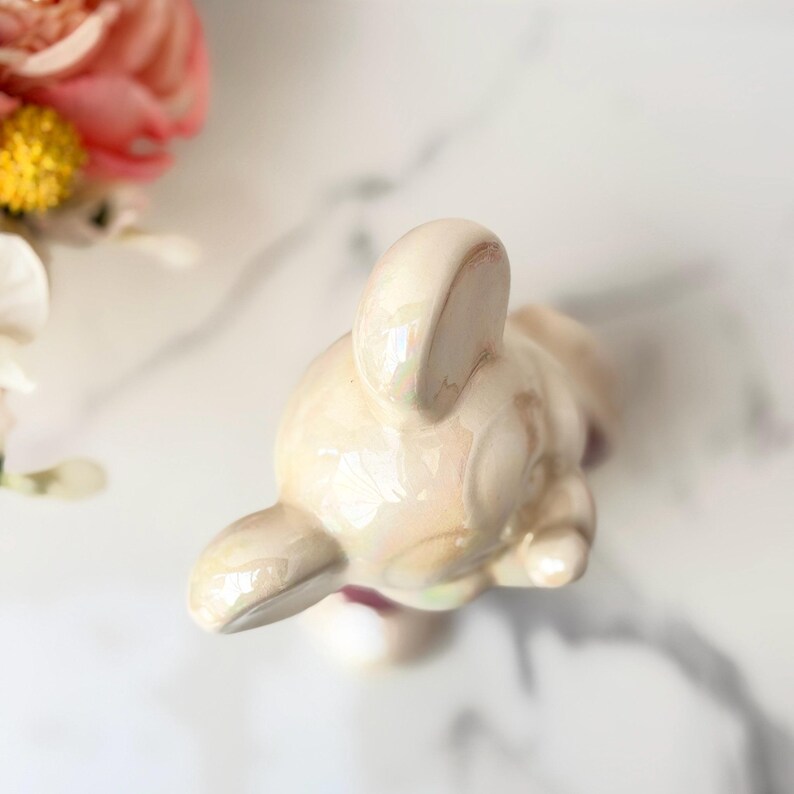 A delicate, iridescent ceramic bunny figurine rests on a white marble surface, with soft pink and yellow flowers blurred in the background.