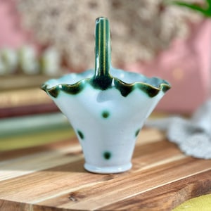 May include: A small, ceramic basket with a handle. The basket is light blue with dark green polka dots and a dark green trim around the scalloped edge. The handle is dark green with vertical lines.