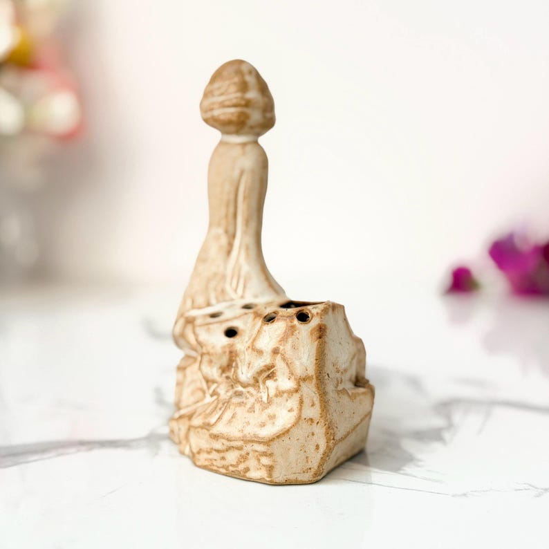 A beige ceramic sculpture of a seated figure on a rock with holes, resembling a pencil sharpener, set against a marble-like surface with pink flowers in the background.
