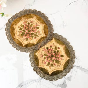 Può includere: Due sottobicchieri circolari in tessuto con un pannello centrale color crema caratterizzato da un motivo floreale ricamato in rosa e verde. I bordi sono rifiniti con un tessuto verde oliva scuro e una frangia decorativa color oro.