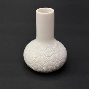 Könnte beinhalten: Eine kleine, weiße Keramikvase mit rundem Boden und langem, zylindrischem Hals. Der Boden ist mit einem erhabenen Blumenmuster verziert. Die Vase ist etwa 15 cm hoch.