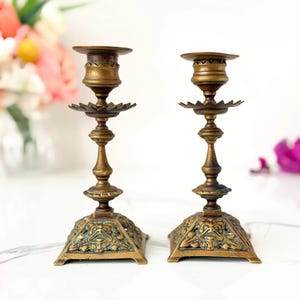 May include: A pair of matching brass candlesticks featuring a square base with intricate relief detailing, a slender central stem with multiple decorative nodes, a scalloped drip plate, and a circular top cup for holding a single taper candle.
