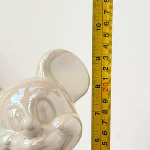 A pearlescent ceramic figurine, possibly a mouse, is measured against a yellow tape measure marked with black numbers from 1 to 9.