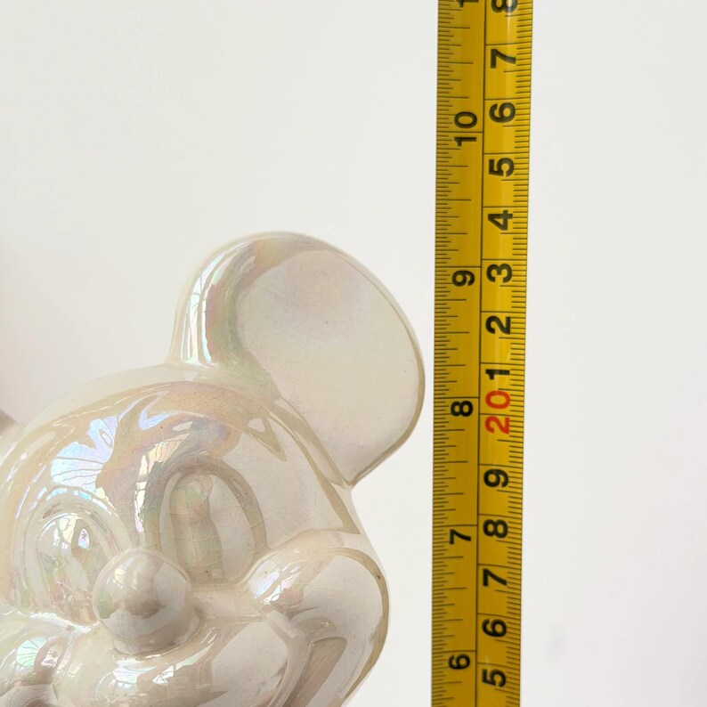 A pearlescent ceramic figurine, possibly a mouse, is measured against a yellow tape measure marked with black numbers from 1 to 9.