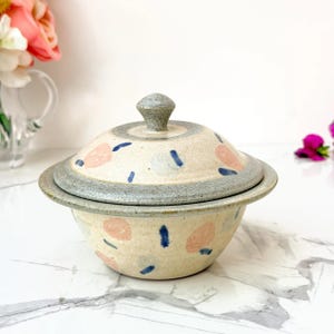May include: A ceramic lidded bowl with a speckled cream base, featuring a hand-painted pattern of soft pink rounded shapes and dark blue dashes. The bowl has a wide rim and a matching lid with a small, rounded knob handle at the center.