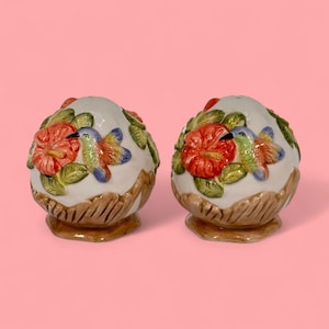 May include: A pair of ceramic salt and pepper shakers. Each shaker is egg-shaped, with a white base and a brown, textured bottom. They are decorated with red hibiscus flowers, green leaves, and a colorful hummingbird.