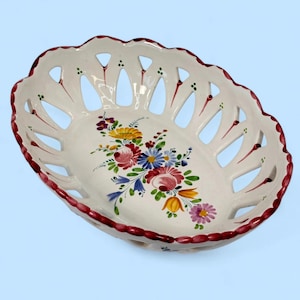 Puede incluir: Cesta ovalada de cerámica blanca con borde festoneado y calado. Decorada con un ramo floral en tonos rosas, azules, amarillos y morados. El borde está rematado con un borde rojo oscuro.