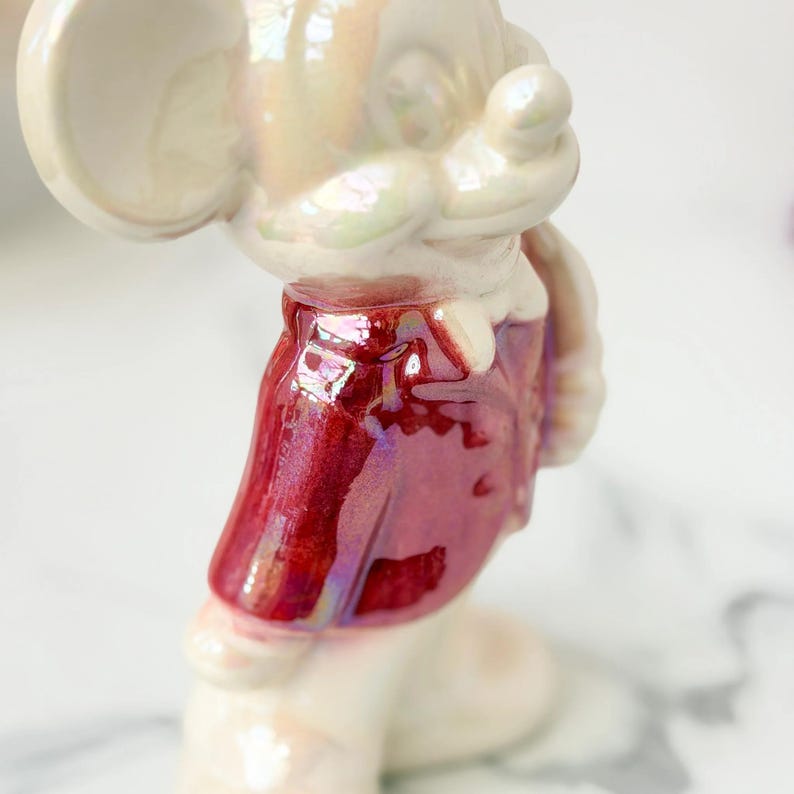 Close-up of a vintage ceramic mouse figurine with an iridescent pearly finish and a sparkly red dress, standing on a white marble surface.