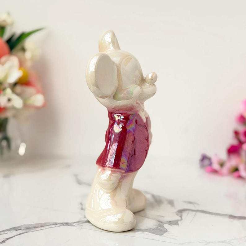 A pearlescent ceramic figurine of Mickey Mouse, wearing a shiny red vest, stands on a marble surface with blurred floral arrangements in the background.