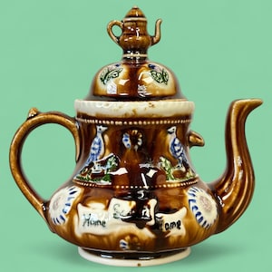 Peut inclure: Théière en céramique marron avec un design décoratif. La théière présente une petite théière sur le couvercle, des oiseaux bleus et les mots "Home Sweet Home". La poignée et le bec sont incurvés.