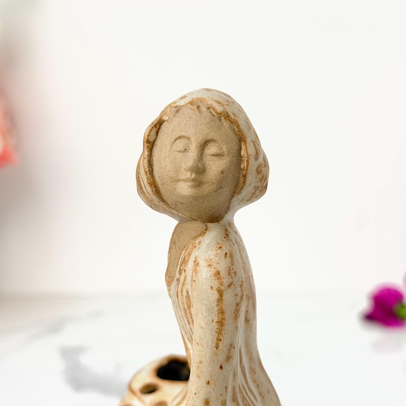 A small, intricately detailed ceramic figurine of a serene woman with closed eyes, wearing a flowing dress, stands gracefully on a white surface.