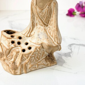 Close-up of a beige ceramic statue with a robed figure, showing multiple circular holes in the chest against a marble surface; purple flowers out of focus in the background.