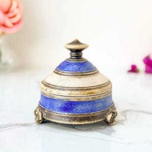 May include: A ceramic lidded jar featuring a rounded base and a pointed finial lid. The design displays alternating bands of deep blue and a cream-colored crackle glaze finish. The base is supported by three small, dark, sculpted feet.