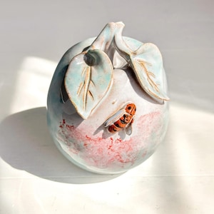 Può includere: Un vaso decorativo in ceramica a forma di pera, con una glassa azzurra e bianca. Due foglie e uno stelo sono in cima. Una piccola ape arancione e nera è attaccata al lato. Il vaso ha un motivo screziato rosso e rosa.