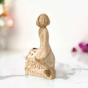 A small ceramic figurine of a girl with closed eyes, sitting on a pedestal with intricate carvings, set on a marble surface.