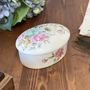 Puede incluir: Una caja de baratijas ovalada, de color blanquecino, con un diseño floral. La tapa presenta un ramo de flores rosas, azules y blancas. Los lados de la caja están adornados con acentos florales similares. La caja está sobre una superficie de madera.