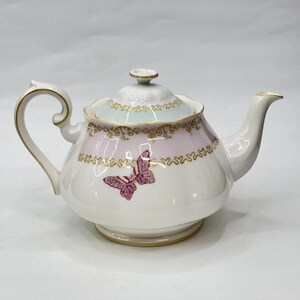Puede incluir: Tetera de porcelana blanca con diseño de mariposa. La tetera presenta una banda rosa y dorada con una ilustración de mariposa morada. La tapa es blanca con un borde azul claro y detalles dorados.