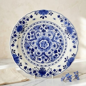 May include: A round ceramic plate with a scalloped edge, featuring a white base decorated with intricate blue floral patterns. The design includes a large central flower surrounded by symmetrical botanical motifs and scrolling vines.