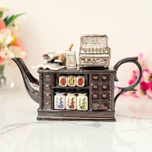 May include: A dark brown ceramic teapot shaped like a vintage shop counter. The top features a miniature cash register, a small book, and a box labeled 'The Tea Shop'. The front displays six small jars with dragon illustrations and the word 'TEA'.