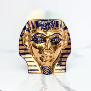 May include: A gold-colored ceramic bust of a pharaoh with blue and gold striped details. The bust features a detailed face with blue eyes and a blue headdress. The interior of the vessel is dark blue.