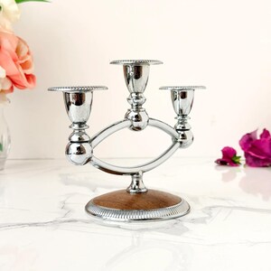 May include: A three-armed silver-toned metal candelabra featuring a central elevated candle cup flanked by two lower cups. The structure is supported by a circular base with a wood-grain inlay and a decorative ridged edge along the bottom rim.