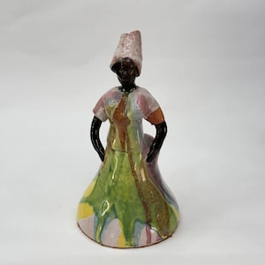 May include: A ceramic figurine of a woman in a colorful dress and head covering. The dress features green, yellow, and pink hues with brown accents. The figure has black arms and a dark face, with hands on hips.