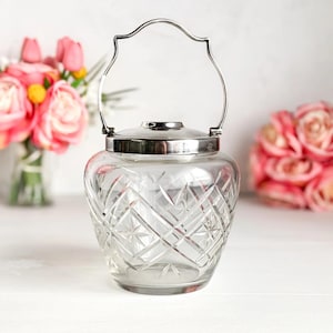 Puede incluir: Un tarro de cristal transparente con tapa y asa plateadas. El tarro tiene un patrón de diamantes y estrellas tallado en el cristal. El asa arqueada está unida a la tapa. El tarro está sobre una superficie blanca.