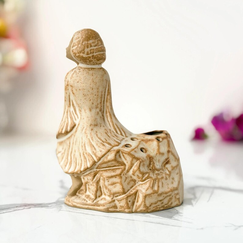 A small terracotta figurine depicts a woman in a flowing dress, seated on a natural rock, with a small hole at the top for a candle, on a marble surface.